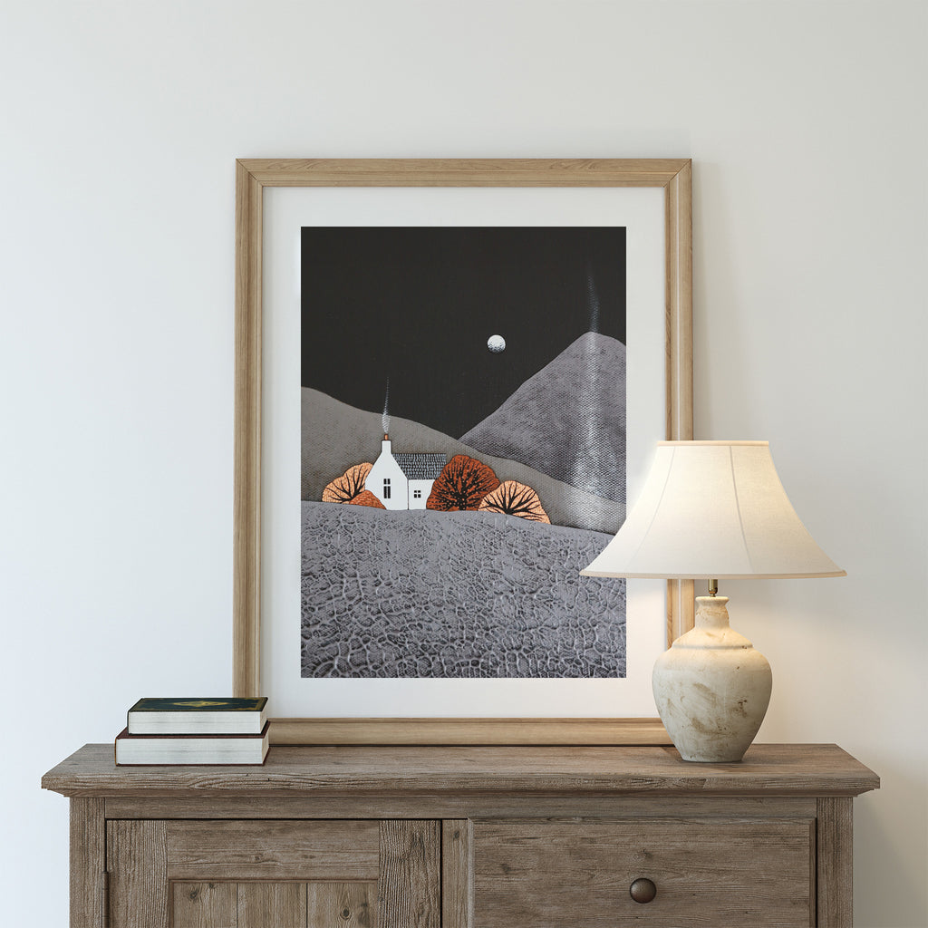 Stunning nature art print featuring a house nestled in an Autumnal landscape. Smoke furls from the chimney into a wintery sky. At print is on a dresser near a lamp and a stack of books.