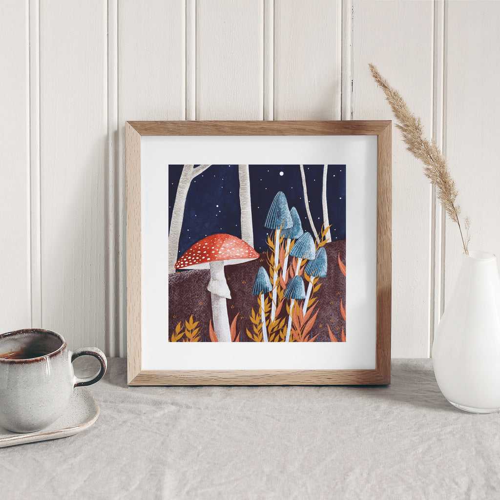 Art print of mushrooms growing in a dark Autumnal forest. Art print is framed and leaning against a white wall.