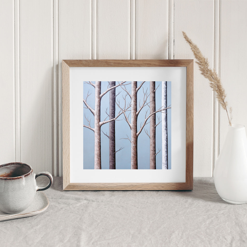 Stunning nature art print featuring a wintery forest, against a brilliant blue sky. Art print is leaning on a table against a wall.
