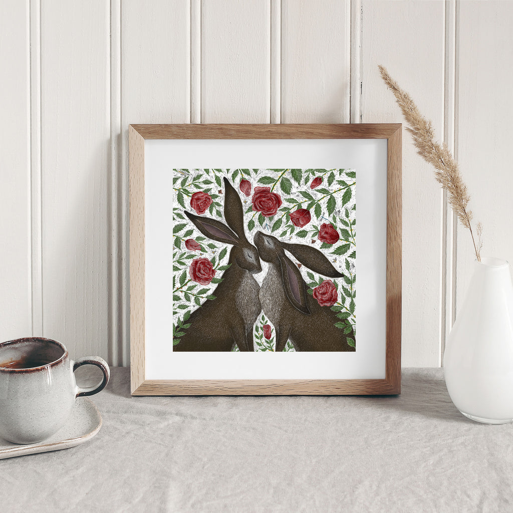 Art print featuring two hares standing in a patch of blooming roses. Art print is leaning on a table against a wall.
