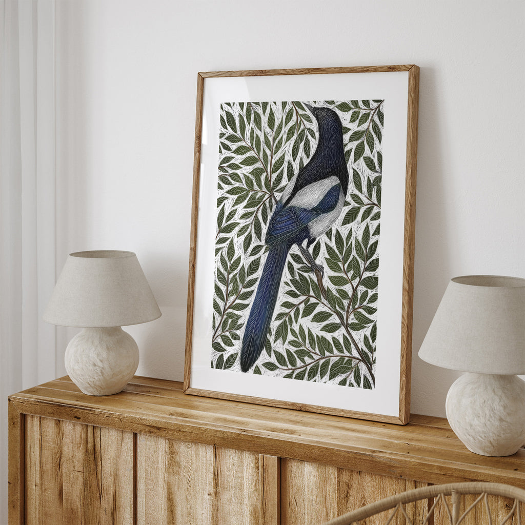Nature art print featuring a detailed illustration of a magpie surrounded by green leaves and branches. Art print is leaning on a table between two lamps.