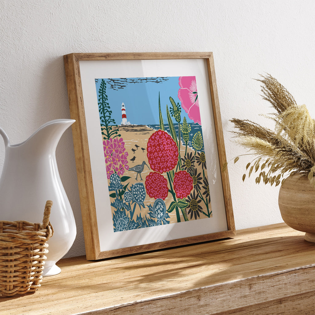 Bright seaside art print featuring flowers, birds and a lighthouse in the distance. Art print is leaning on a shelf near dining room items.