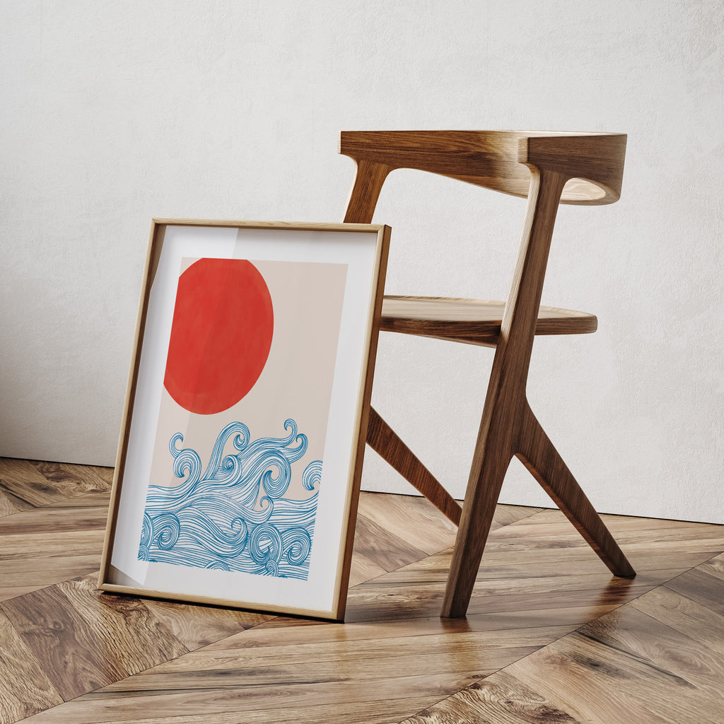 Beautiful art print featuring waves reaching up to a red sun. Art print is framed and leaning against a wooden chair.