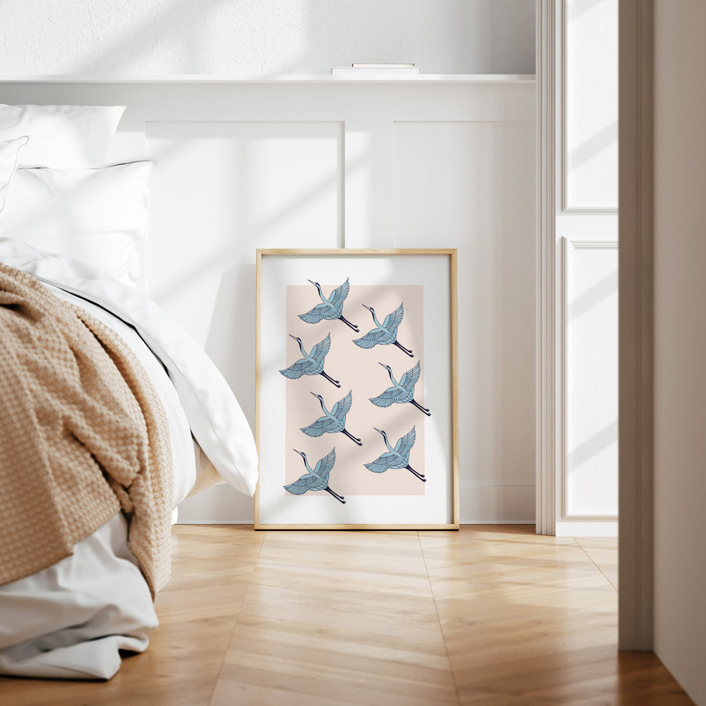 Detailed art print featuring a pattern of heron's flying in formation, on a pale pink background. Art print is leaning against a panelled wall next to a bed.