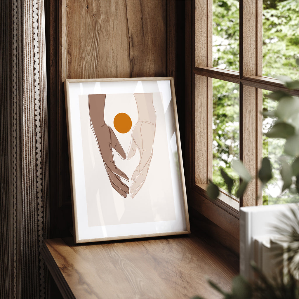 Beautiful art print featuring two hands meeting in the meeting of the frame. A bright sun sits behind the hands. Art print is framed and sits on a window ledge. 
