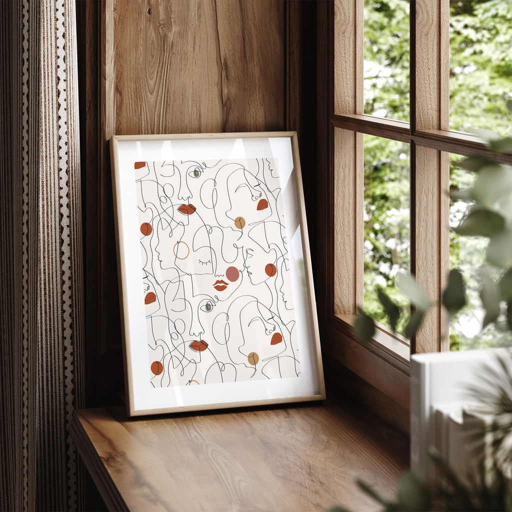 Pastel art print featuring clever line work to create a pleasing pattern of faces. Pink and beige colours are used to emphasise lips and cheeks. Art print is leaning on a window seat.