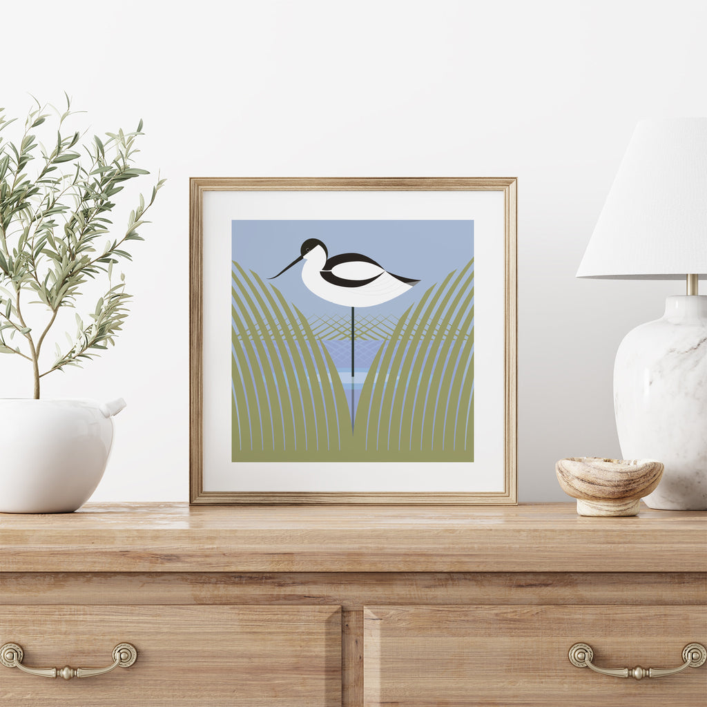 Stuart Cox art print featuring an avocet. Drawn in pastel colours using geometric shapes. Art print is stood on a dresser between a plant and lamp.