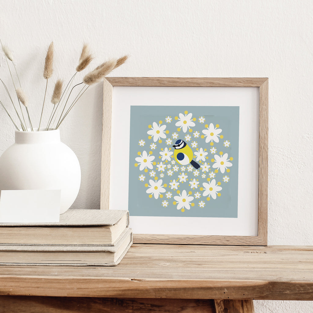 Minimalistic art print featuring a beautiful blue tit surrounded by bright Spring flowers. Art print is leaning against a white wall.