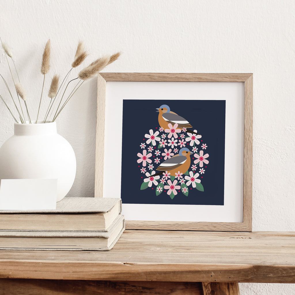 A striking geometric art print of two birds amidst a blooming circlet of flowers. Art print is on a table with some books.