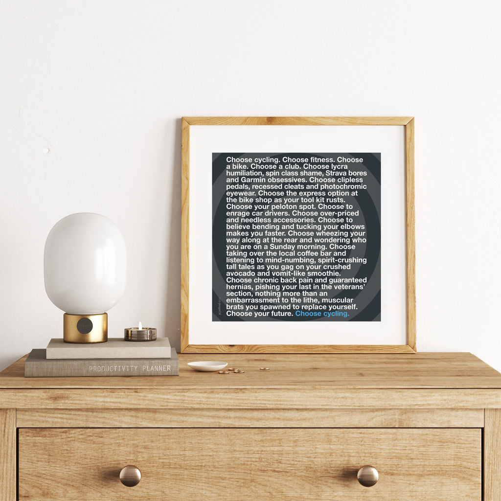 Sporty art print featuring a long paragraph of typography celebrating cycling, on a black background. Art print is leaning on a chest of drawers.