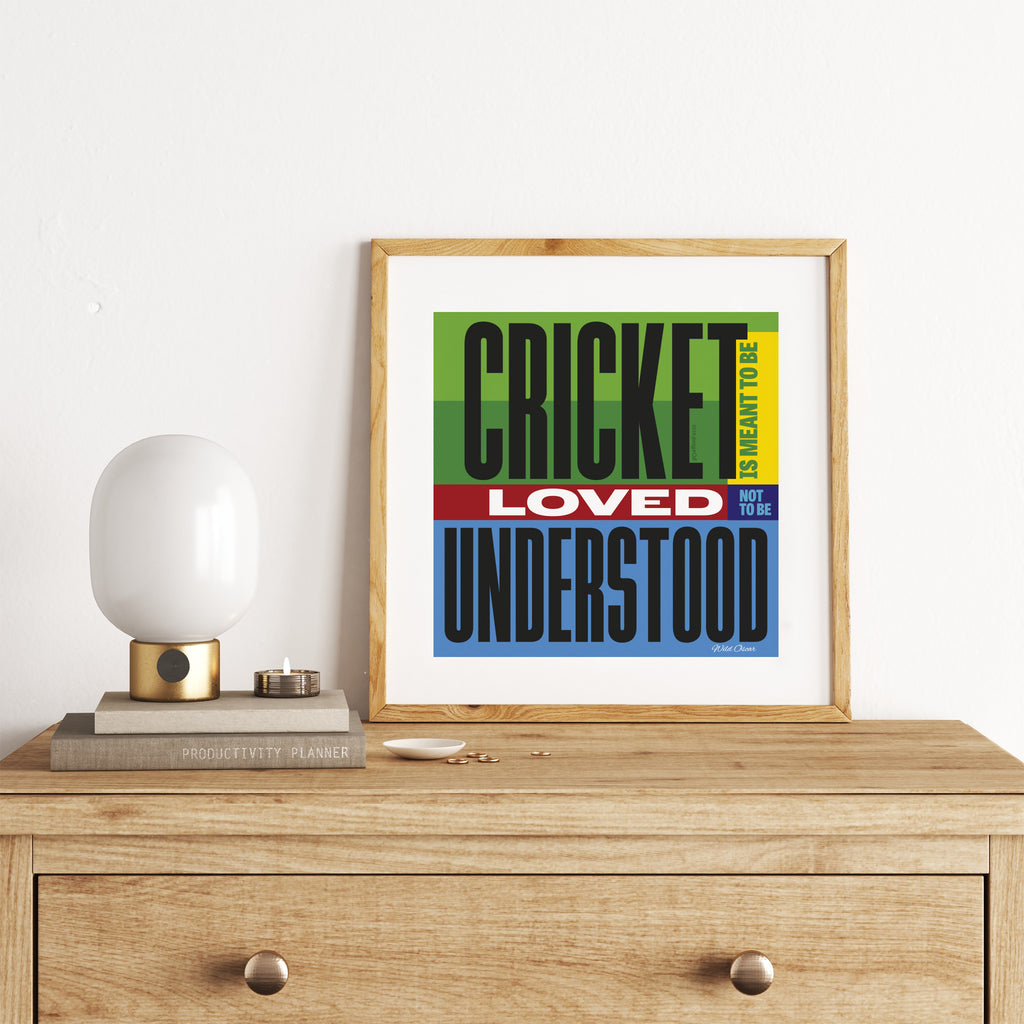 Eccentric art print featuring text and colour, celebrating cricket. Text spells out 'Cricket is meant to be loved, not to be understood'. Art print is stood on a chest of drawers next to a lamp.