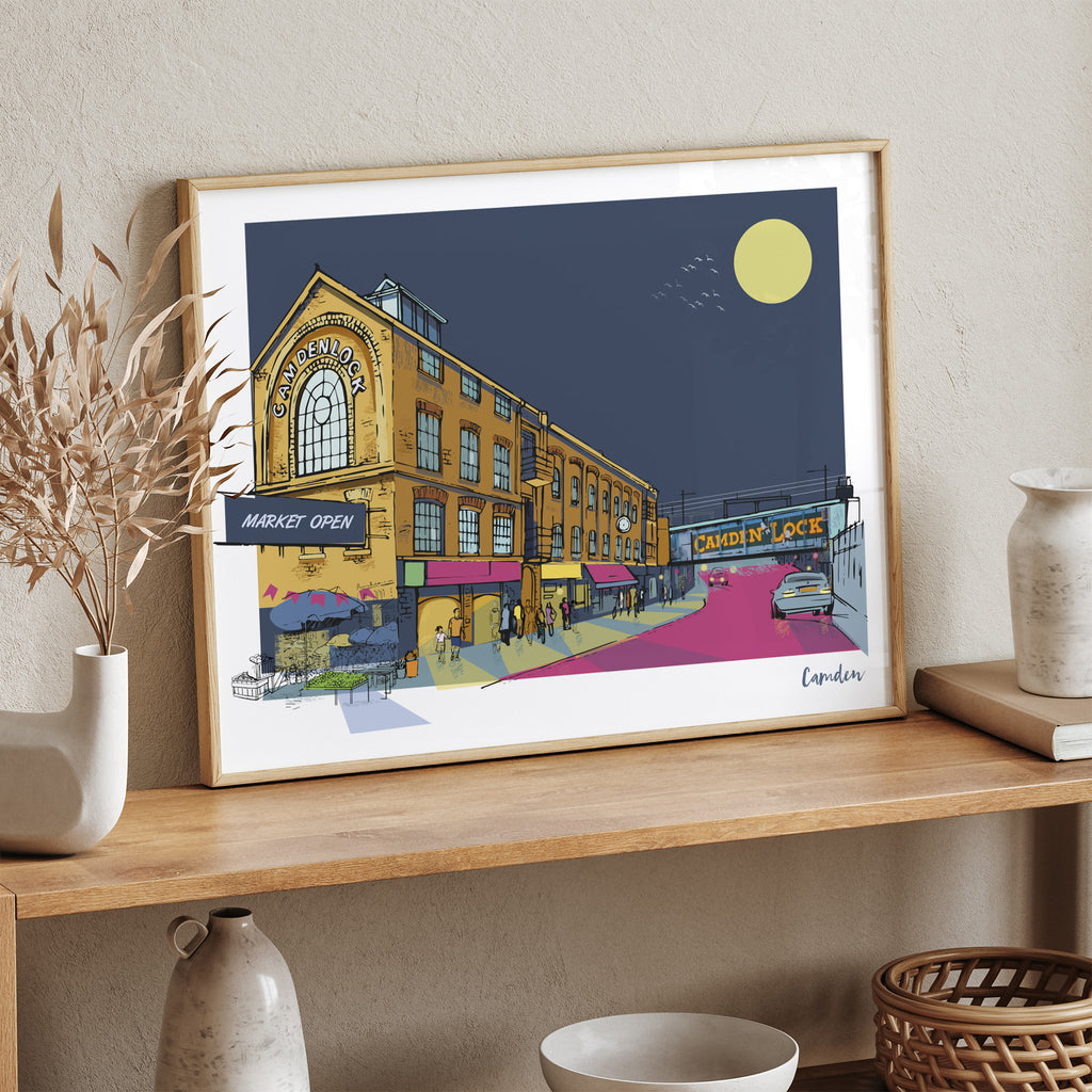Travel art print of Camden Market, London, at night, featuring bright and moody colours, with the title 'Camden' in the bottom right. Art print is leaning on a table against a wall.