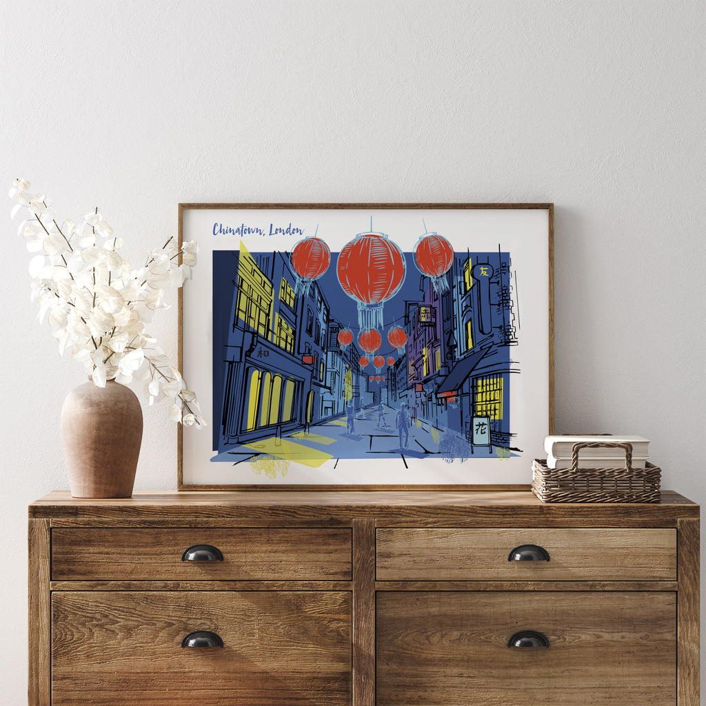 London travel art print featuring a moonlit Chinatown in London, with iconic red lanterns featured against a deep blue background, with the title 'Chinatown, London' in the top left. Art print is stood on a chest of drawers, leaning against a white wall.