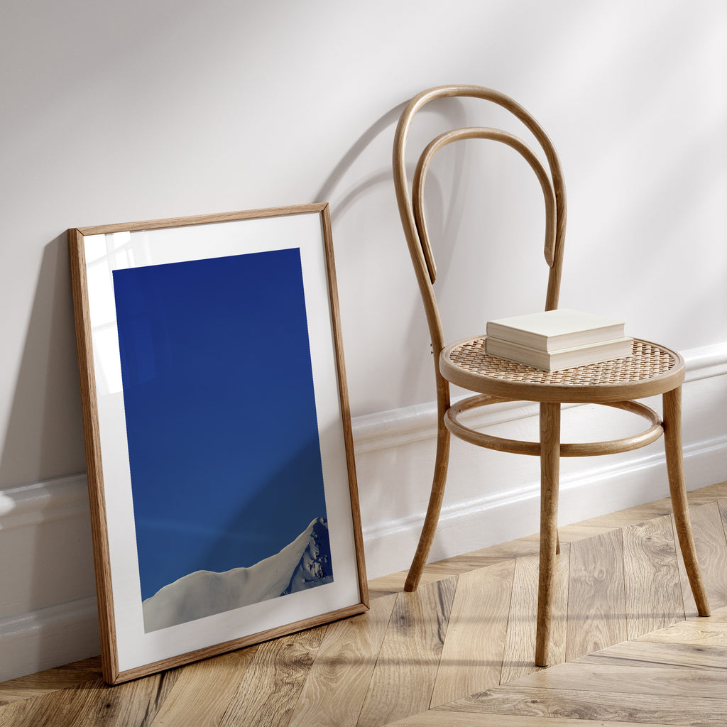 Photography art print featuring a snowy peak of Beinn Eighe, under a brilliant blue sky. Art Print is leaning against a white wall next to a desk chair.