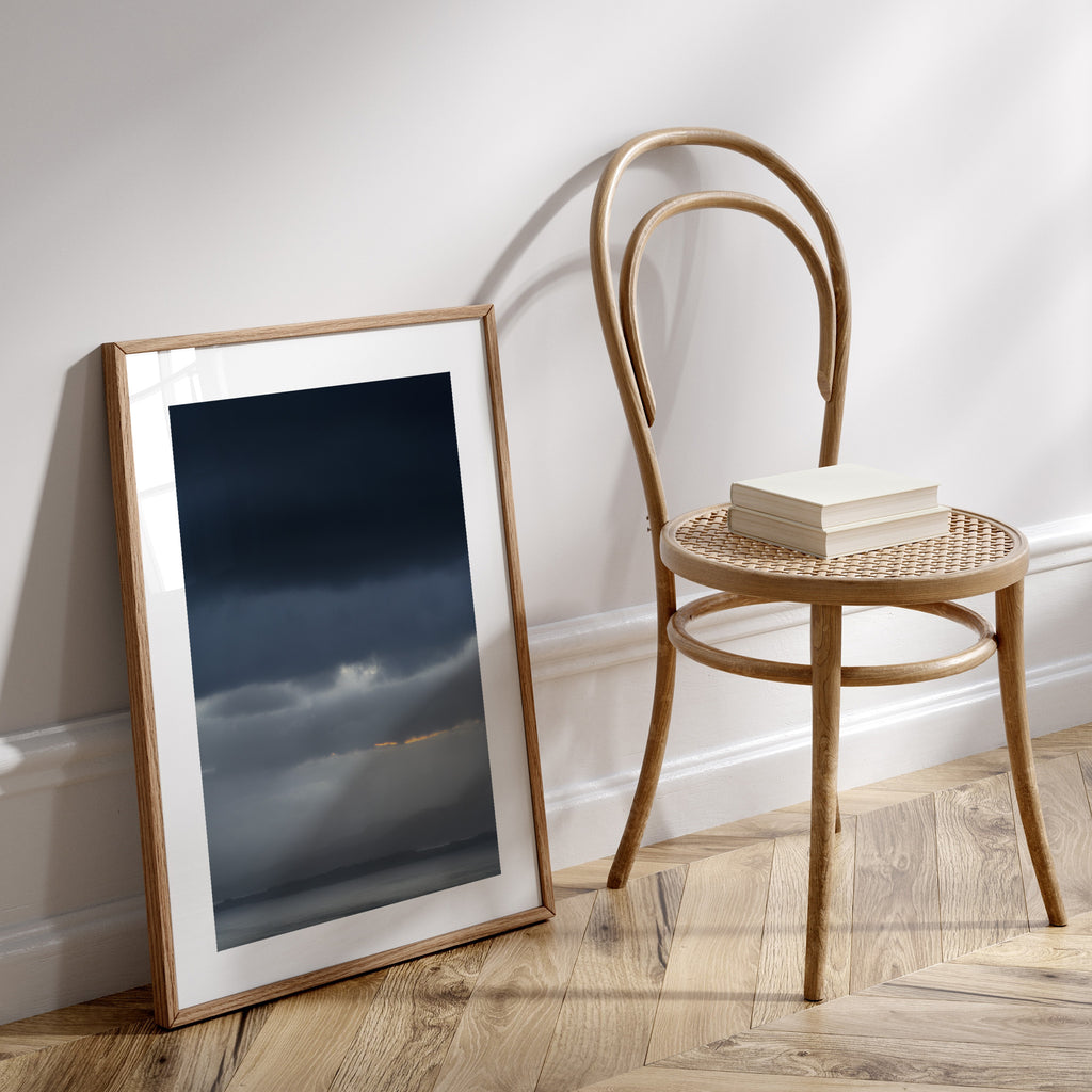 Photography art print featuring a vivid, cloudy sunrise over moody waters. Art print is leaning on the floor against a white wall next to a chair.