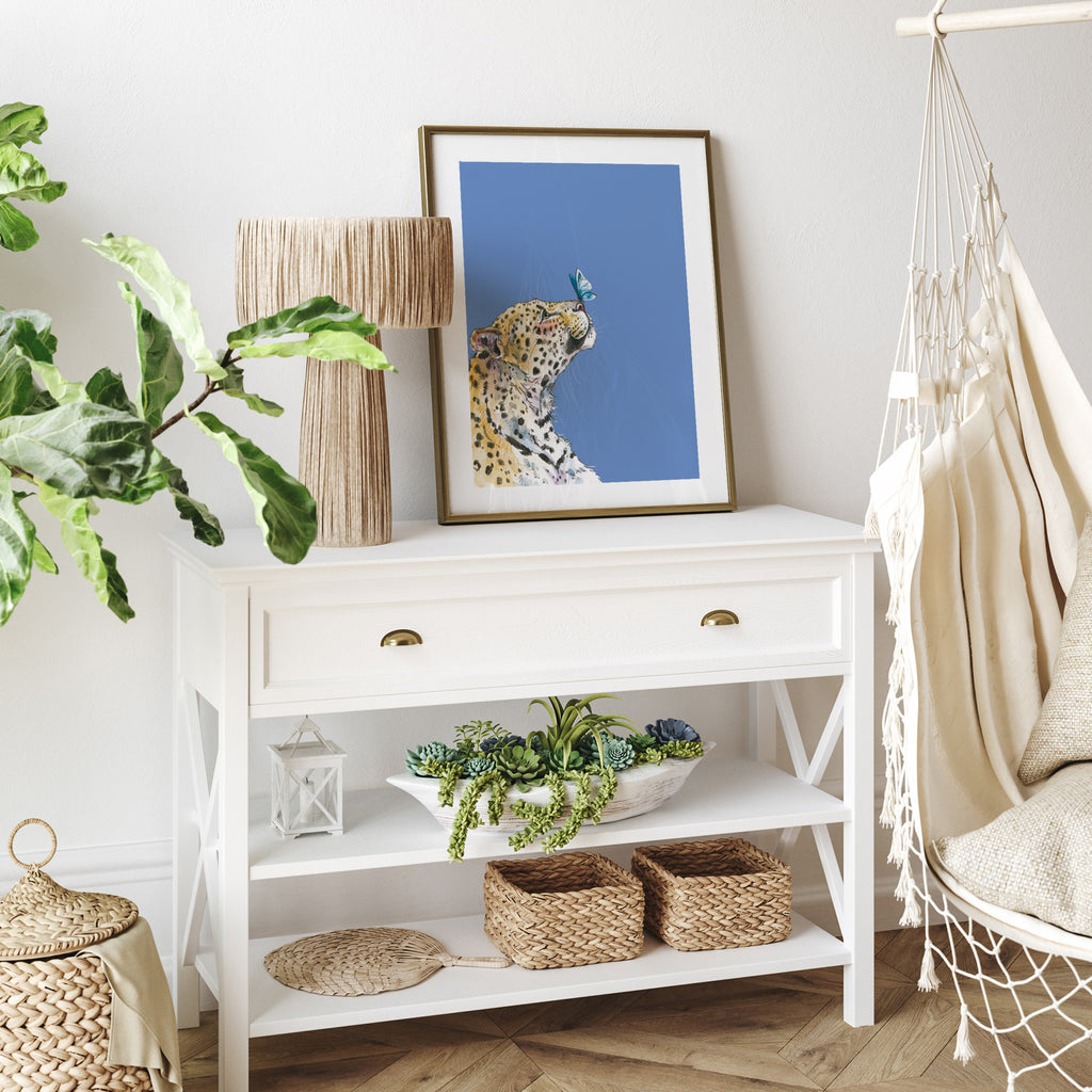 Animal art print featuring a cheetah gazing up at a butterfly on its nose, in front of a pastel blue background. Art print is on a table near lots of plants and a hammock.
