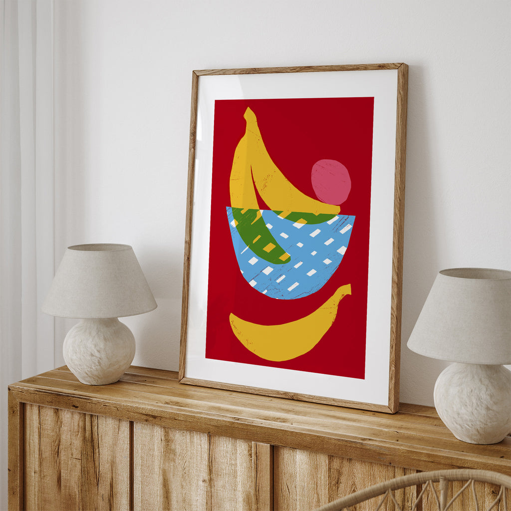 Vivid art print containing bananas playfully placed around a fruit bowl, with a red background. Art print is leaning on a sideboard next to two lamps.
