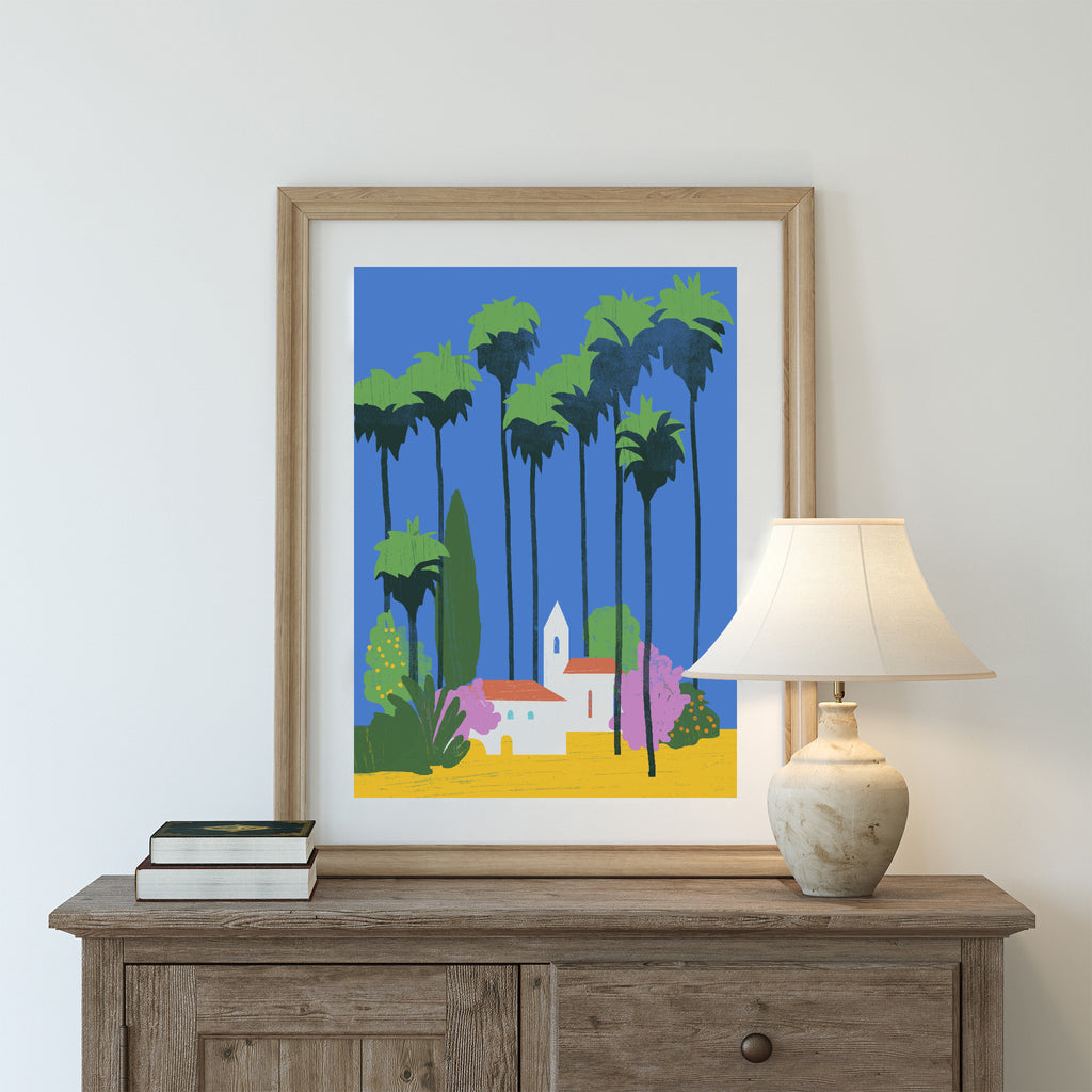 Bright art print featuring a beautiful tropical scene of houses under sunny palm trees. Art print is on a chest of drawers next to a lamp.