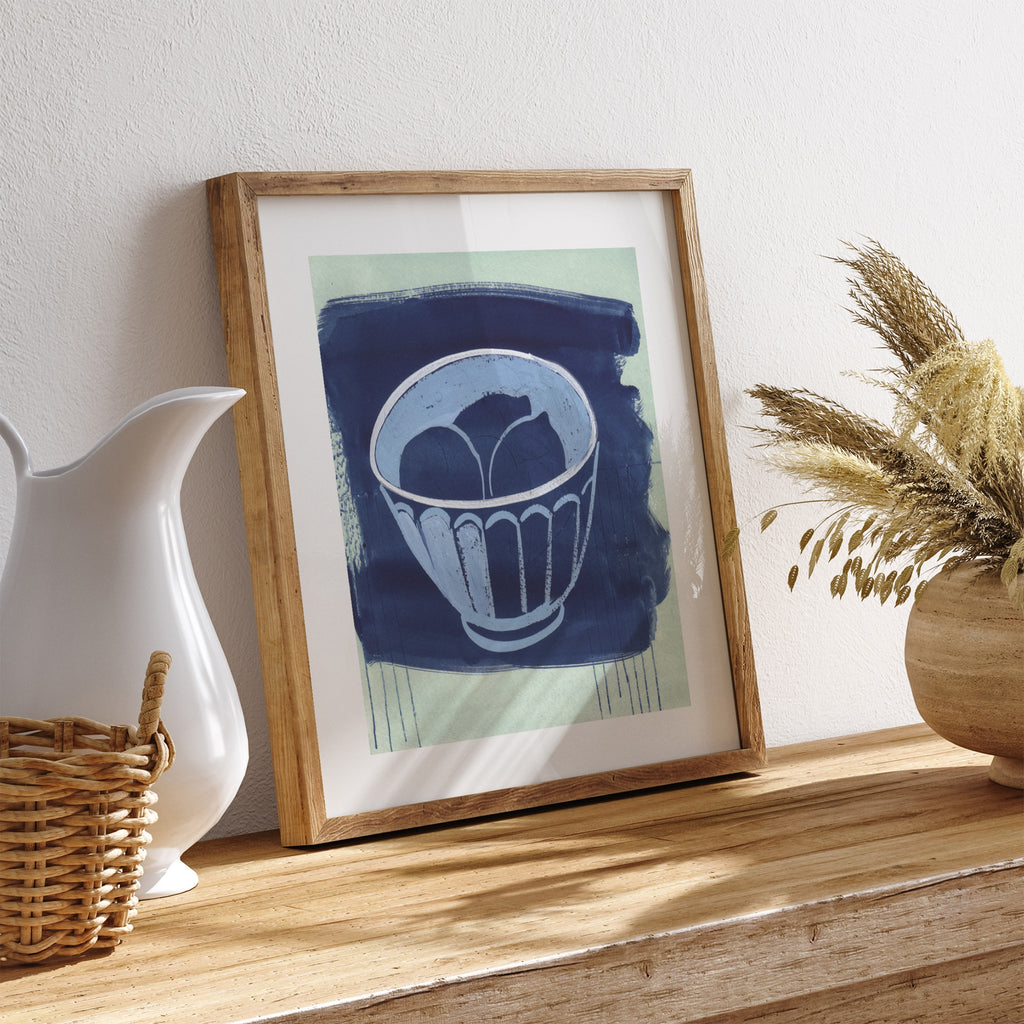 Art print of a bowl of fruit, on a blue background. Art print is leaning on a table with a basket, jug and plant nearby.