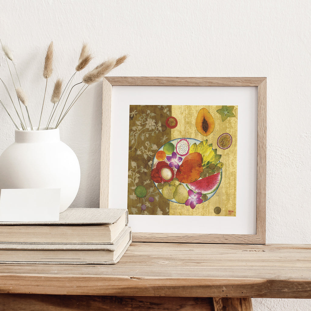 Fruity art print featuring a vibrant fruit platter displayed with flowers on a yellow and brown background. Art print is stood on a dresser next to some books.