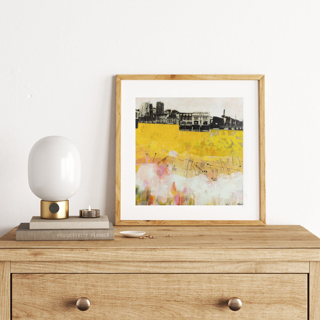 Abstract art print featuring an urban landscape standing on a bright yellow map. Art print is leaning on a chest of a drawers against a wall.