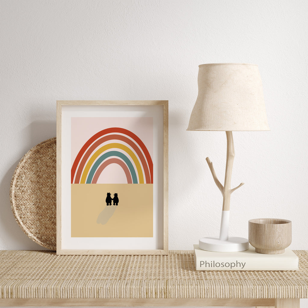 'Bear' art print of two bears gazing up at a large rainbow. Art print is leaning against a. white wall next to a wooden lamp.