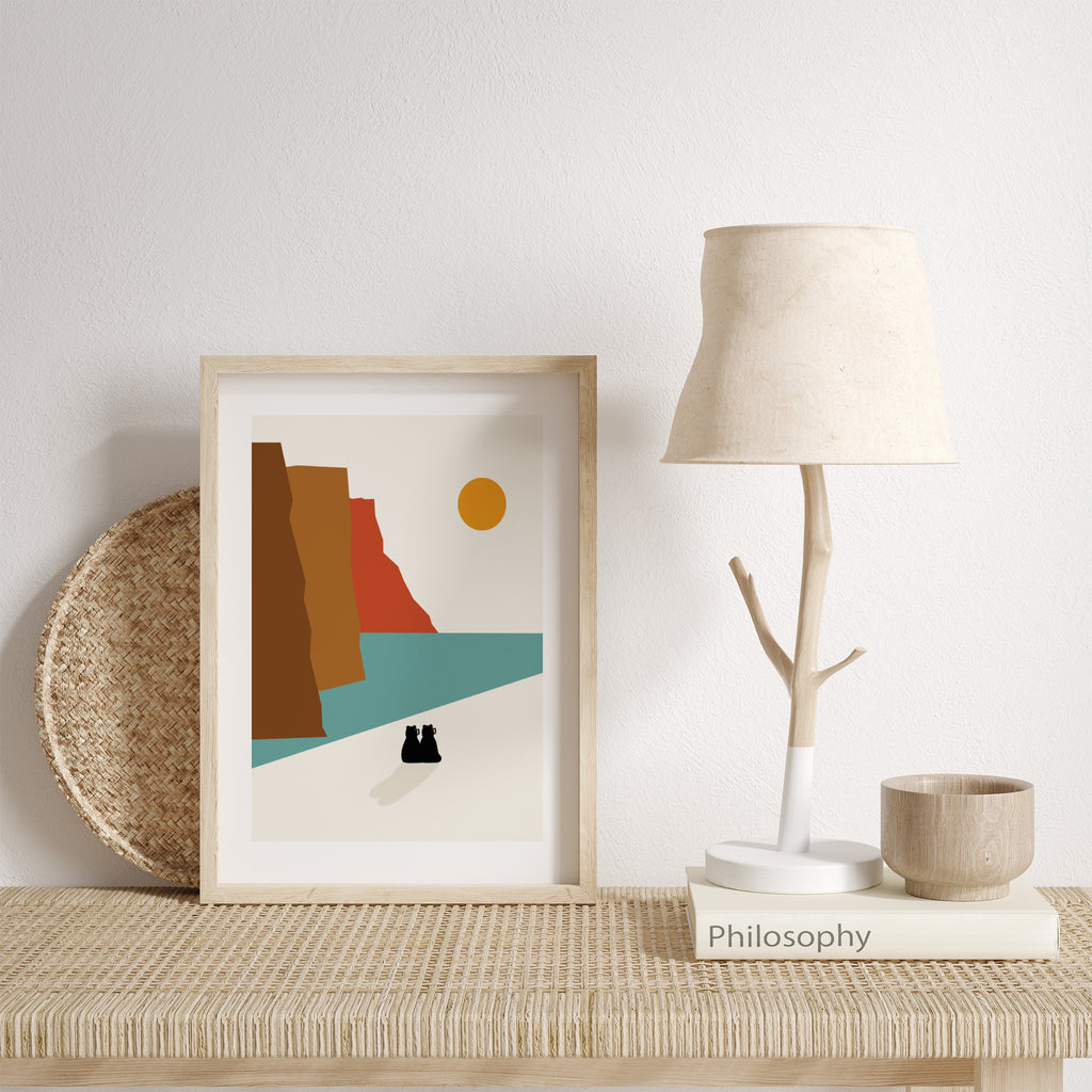 Colourful pastel art print featuring two bears sat watching the scenery before them. Three cliffs stand next to a bright blue ocean, where an orange sun sits in the sky. Art print is leaning against a white wall next to a playful lamp.