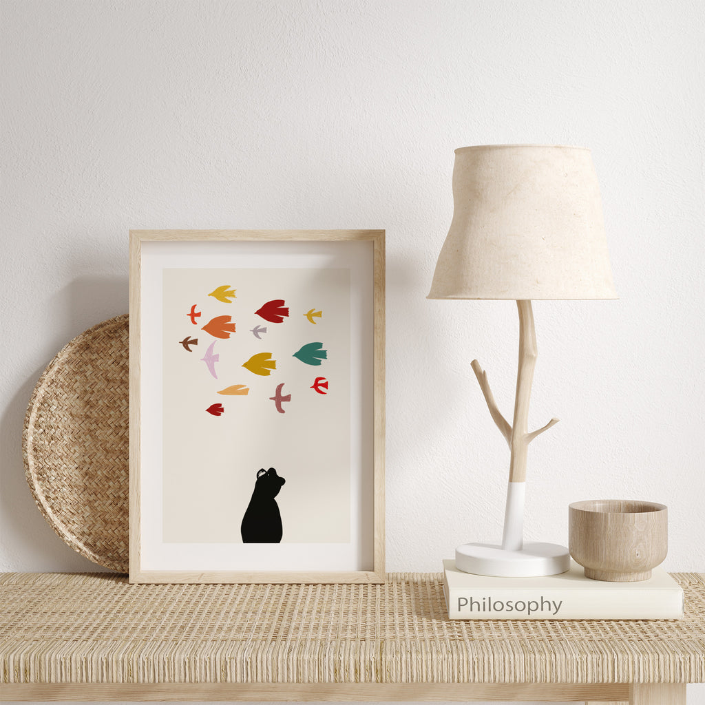 'Bear' art print of bear gazing up at a vibrant flock of birds passing by. Art print on a table leaning against a wall.