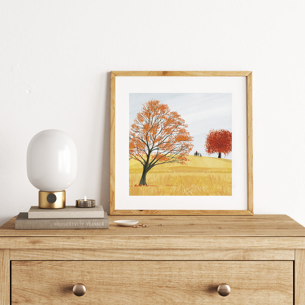 Autumn art print featuring vivid red and orange trees and grass, based off of Richmond Park, London. Someone sits on a bench in the distance, with their dog. Art print is leaning on a chest of drawers against a white wall.