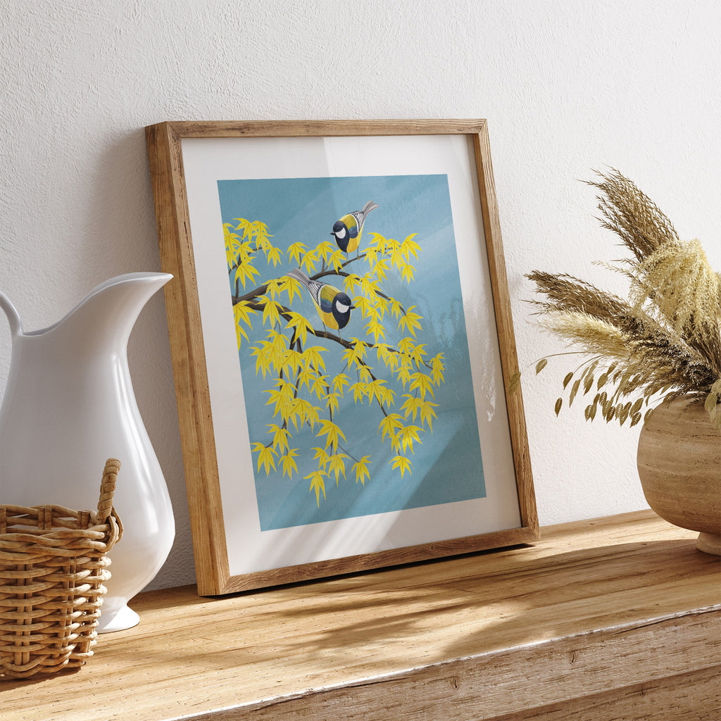 Nature art print featuring two blue tits standing on a malting Autumn tree, in front of a brilliant blue sky. Art print is leaning against a wall on a chest of drawers.