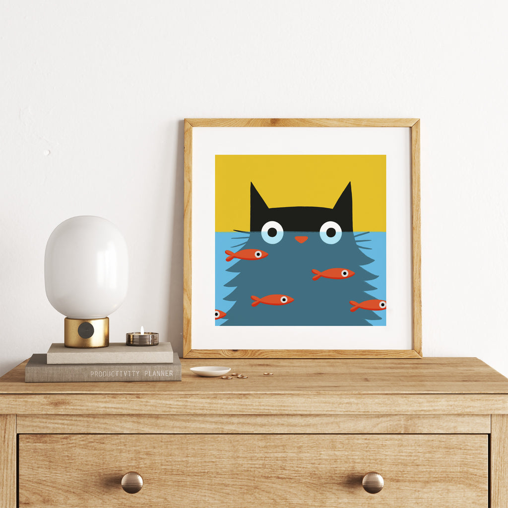 Bright art print featuring a black cat stood in front of a goldfish bowl. Art print is stood on a dresser.