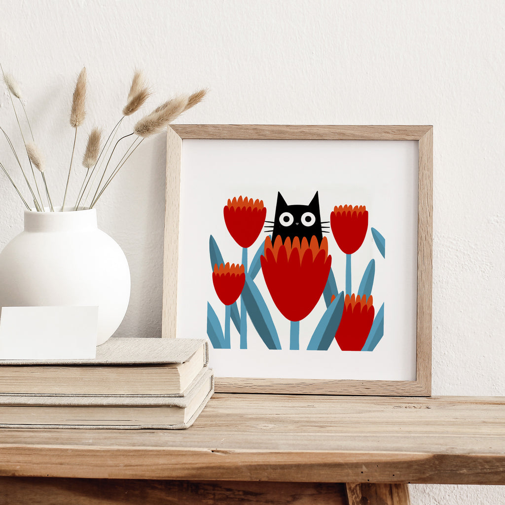 Fun art print of a small black cat posing in a patch of red flowers. Art print is leaning against a wall next to a stack of books.