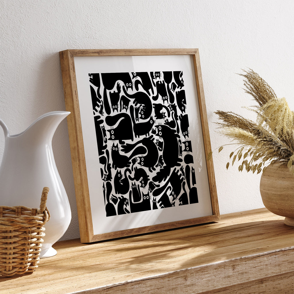 Playful art print of a pattern of many black cats. Art print is leaning against a white wall on a table.