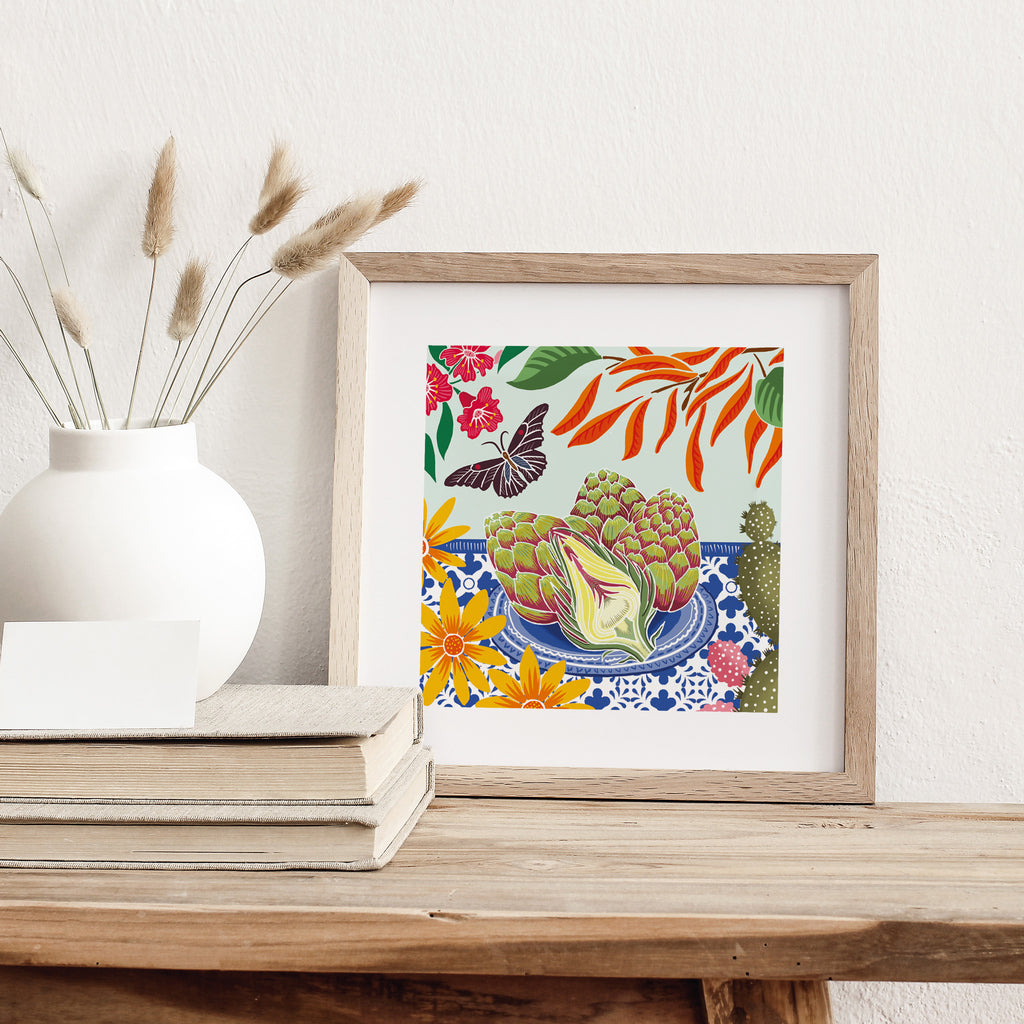 Art print of artichokes on a floral table surrounded by flowers and a butterfly. Art print is leaning against a wall on a table.
