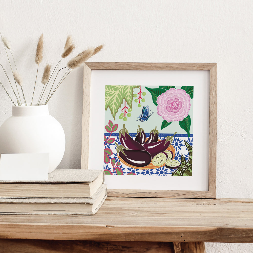 Art print of a plate of an aubergines surrounded by flowers and a butterfly. Art print is leaning against a wall on a table.