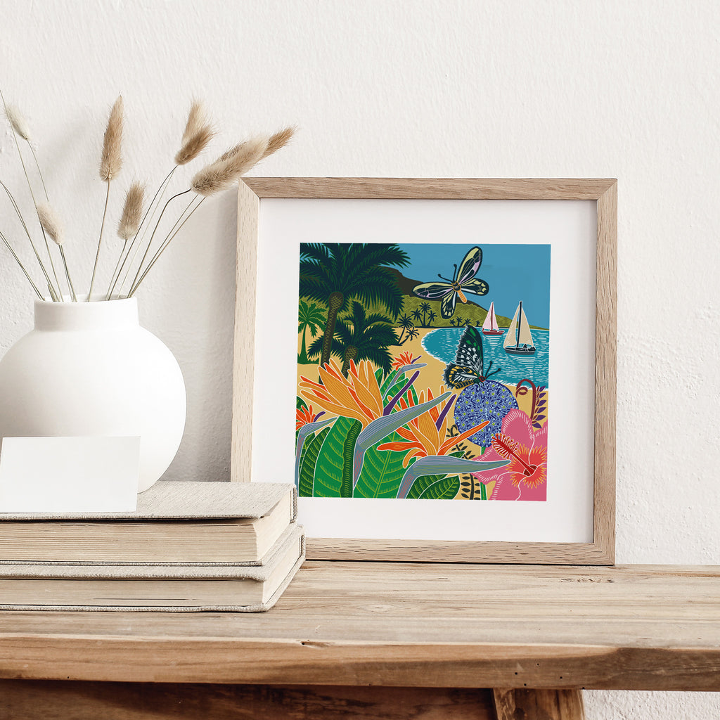 Art print featuring butterflies flitting over a tropical beach with sailboats in the bay. Art print is leaning against a white wall.