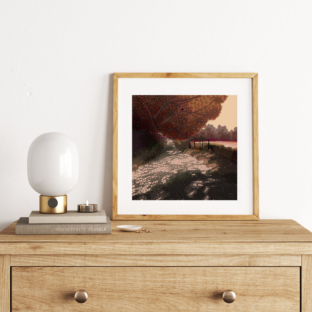Autumnal art print of a path surrounded by red and orange trees and foliage. Art print is leaning against a white wall on a dresser.