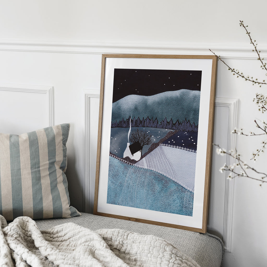 Art print featuring a cosy home in blue midnight countryside fields. Art print is leaning on a sofa in a living room. 