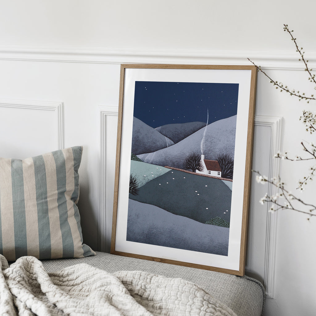 Art print featuring a cosy home nestled in rolling midnight hills in the evening countryside. Art print is leaning on a sofa in a living room.