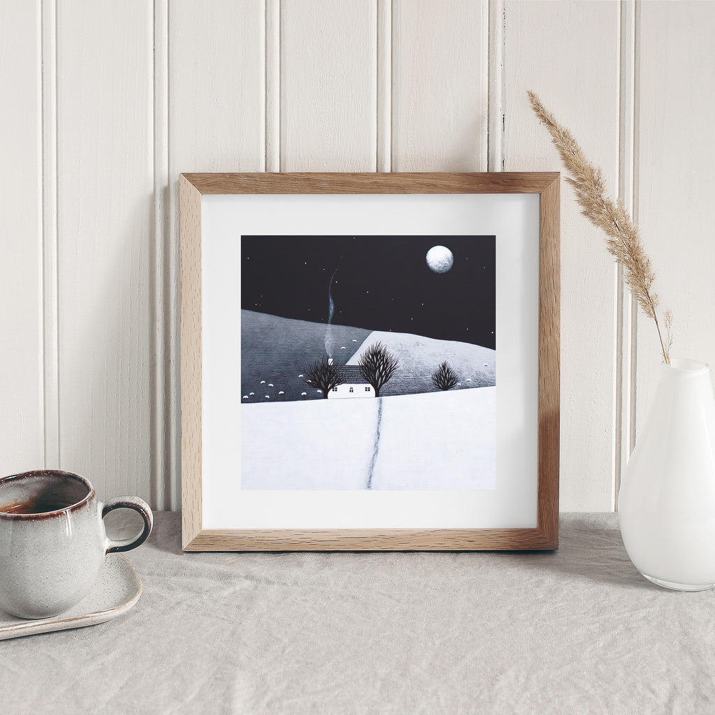 Art print of a cosy home in a wintery moonlit field. Art print is leaning against a white wall.