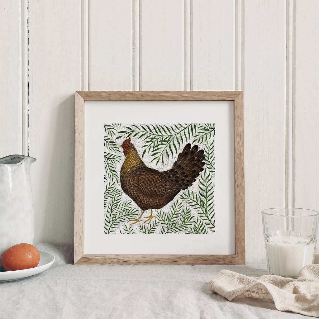 Art print featuring an illustration of a chicken surrounded by green leaves. Art print is leaning on a kitchen table next to eggs and milk.