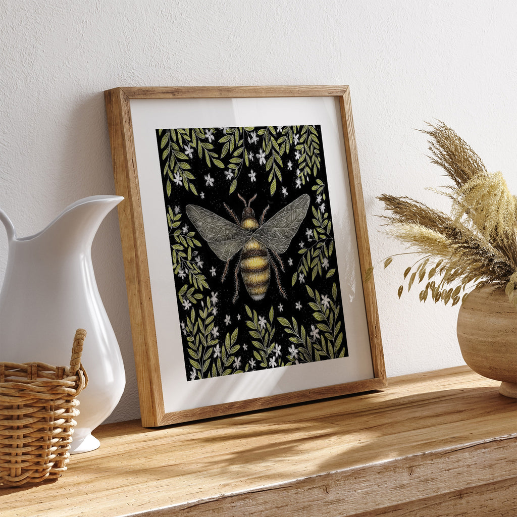 Art print featuring an English bumblebee surrounded by green leaves and white flowers on a black background. Art print is leaning on a shelf against a white wall.