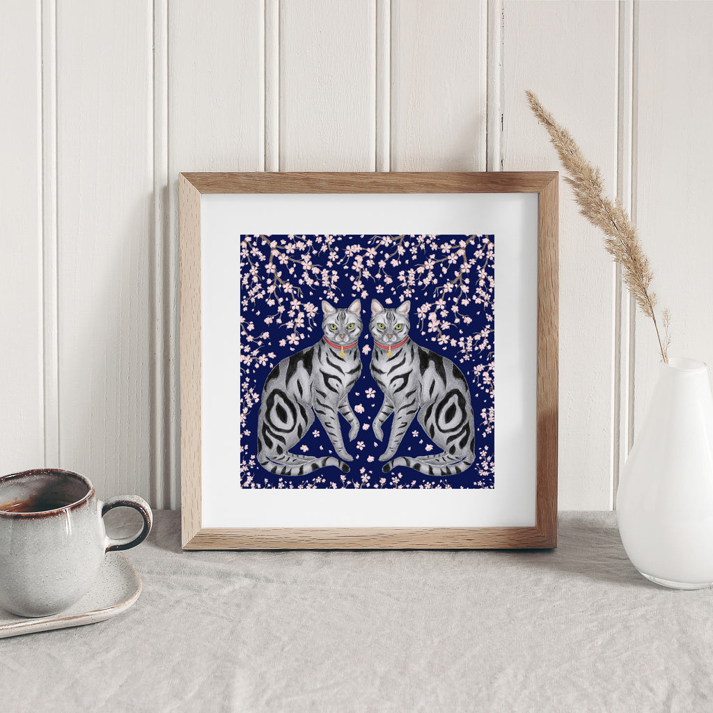 Art print of symmetrical striped cats surrounded by sakura blossoms and a deep blue background. Art print is stood next to kitchen items and leaning against a wall.