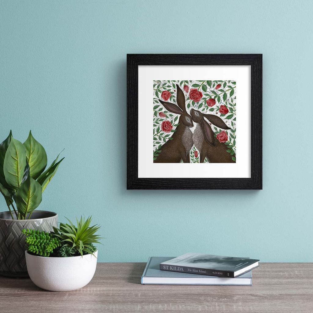 art print features two hares standing in a patch of blooming roses.  Art print is hung up on a blue wall.