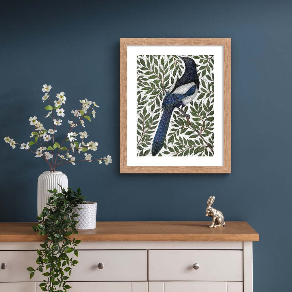 Nature art print featuring a detailed illustration of a magpie surrounded by green leaves and branches. Art print is hung up on a dark blue wall.