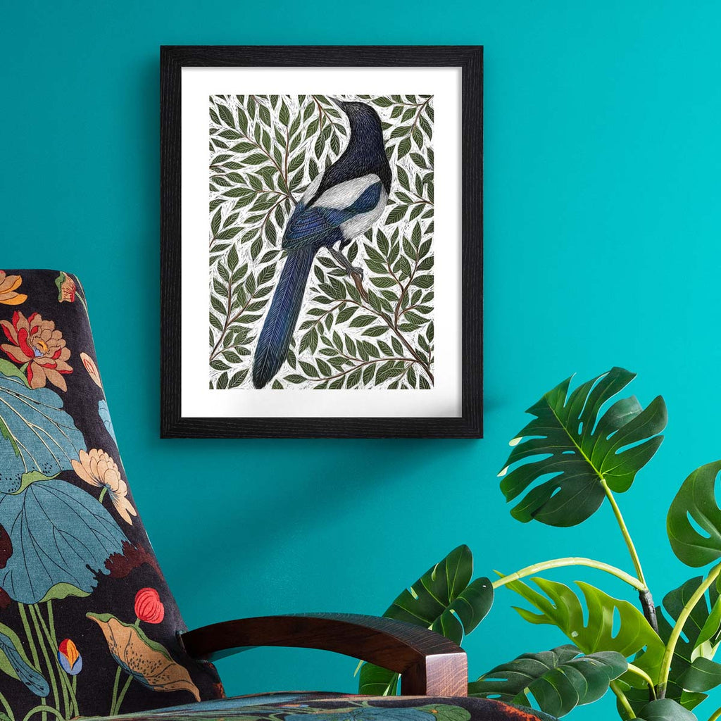 Nature art print featuring a detailed illustration of a magpie surrounded by green leaves and branches. Art print is hung up on a bright blue wall.
