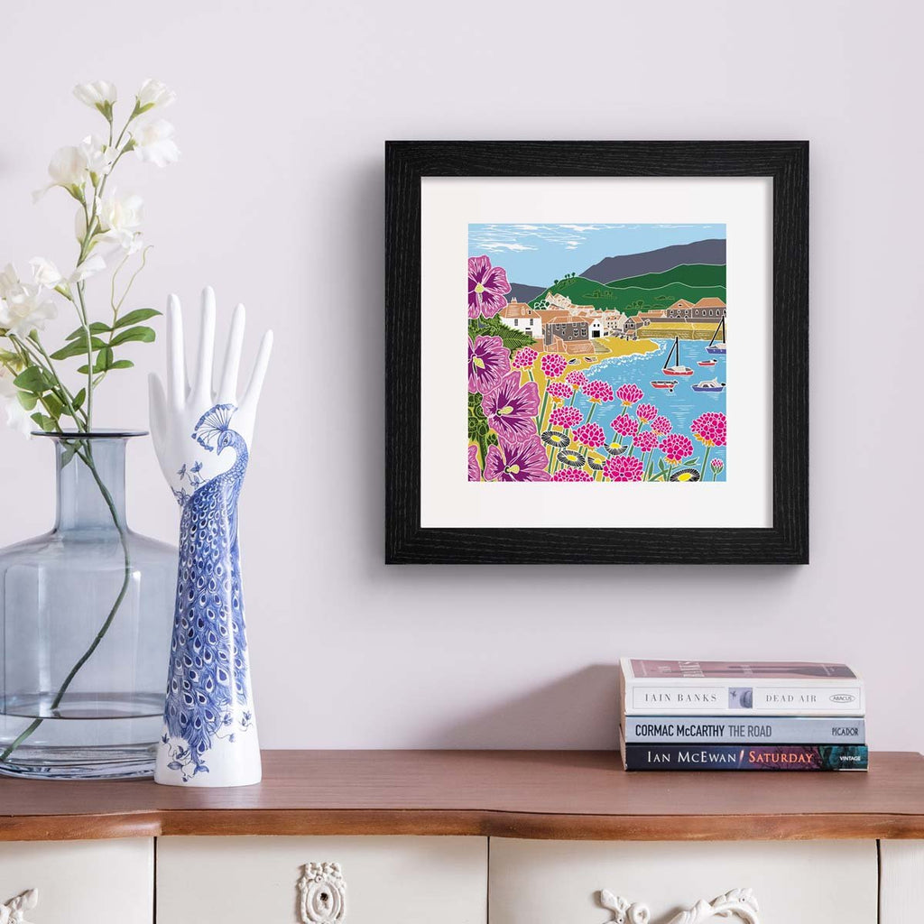 Colourful nature art print featuring a coastal scene of a seaside town, the beach and blooming botanicals. Art print is hung up on a white wall.