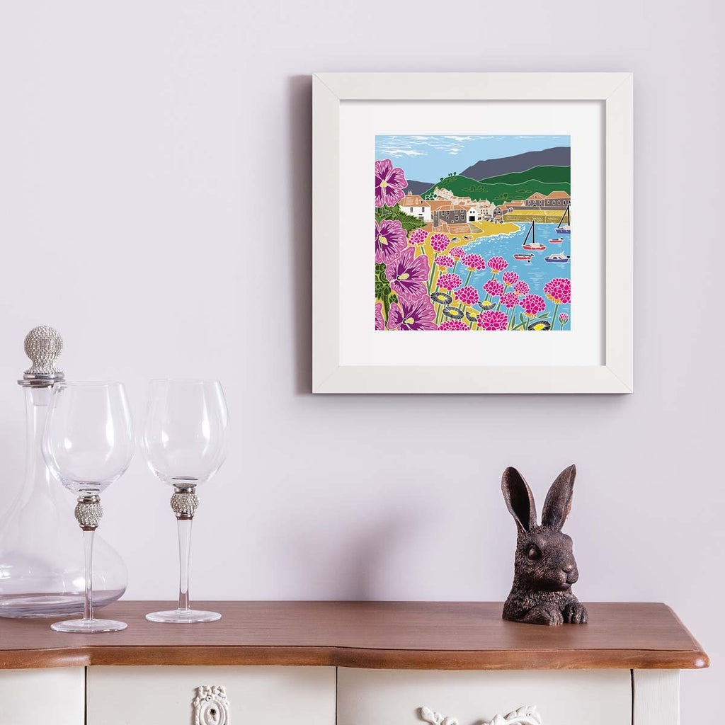 Colourful nature art print featuring a coastal scene of a seaside town, the beach and blooming botanicals. Art print is hung up on a pale pink wall.