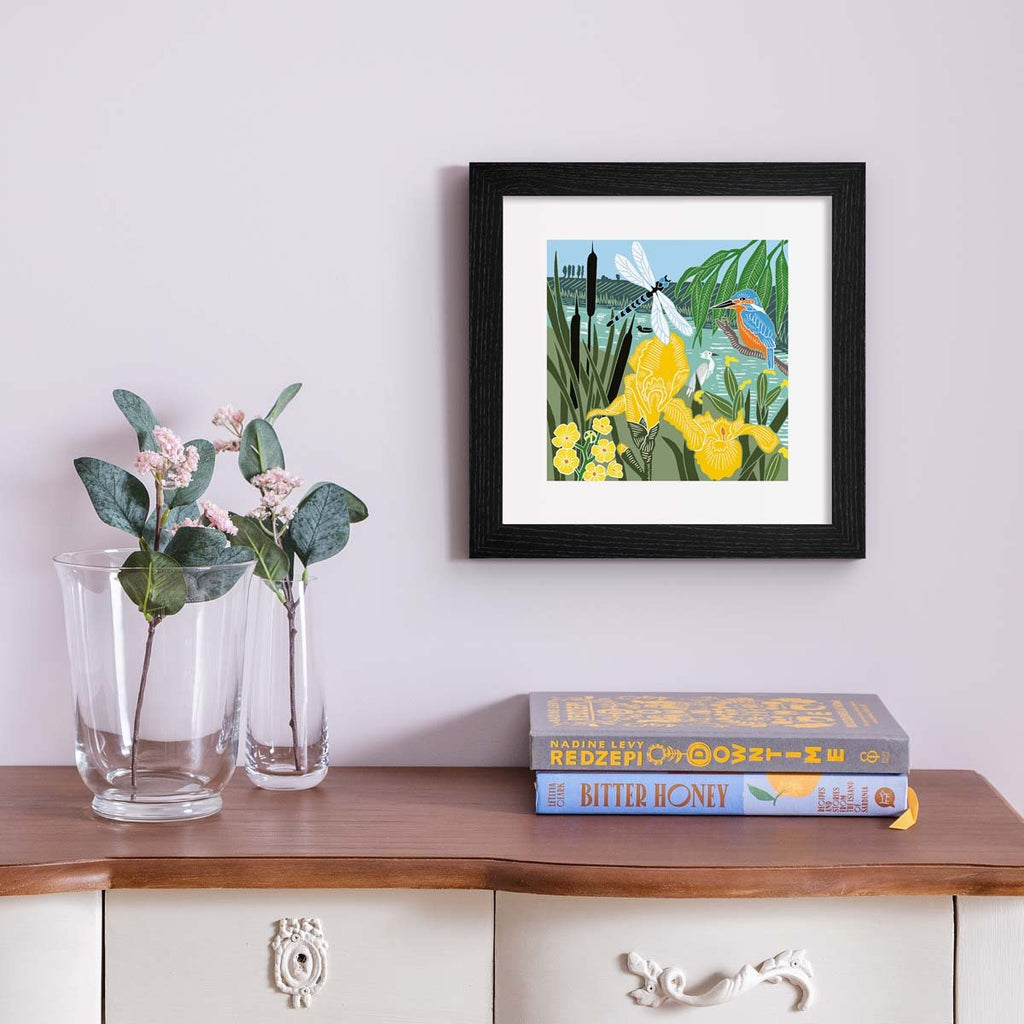 Beautiful art print containing a picturesque Summer waterside scene, featuring animals and botanicals frolicking in the greenery. Art print is hung up on a pale pink wall.