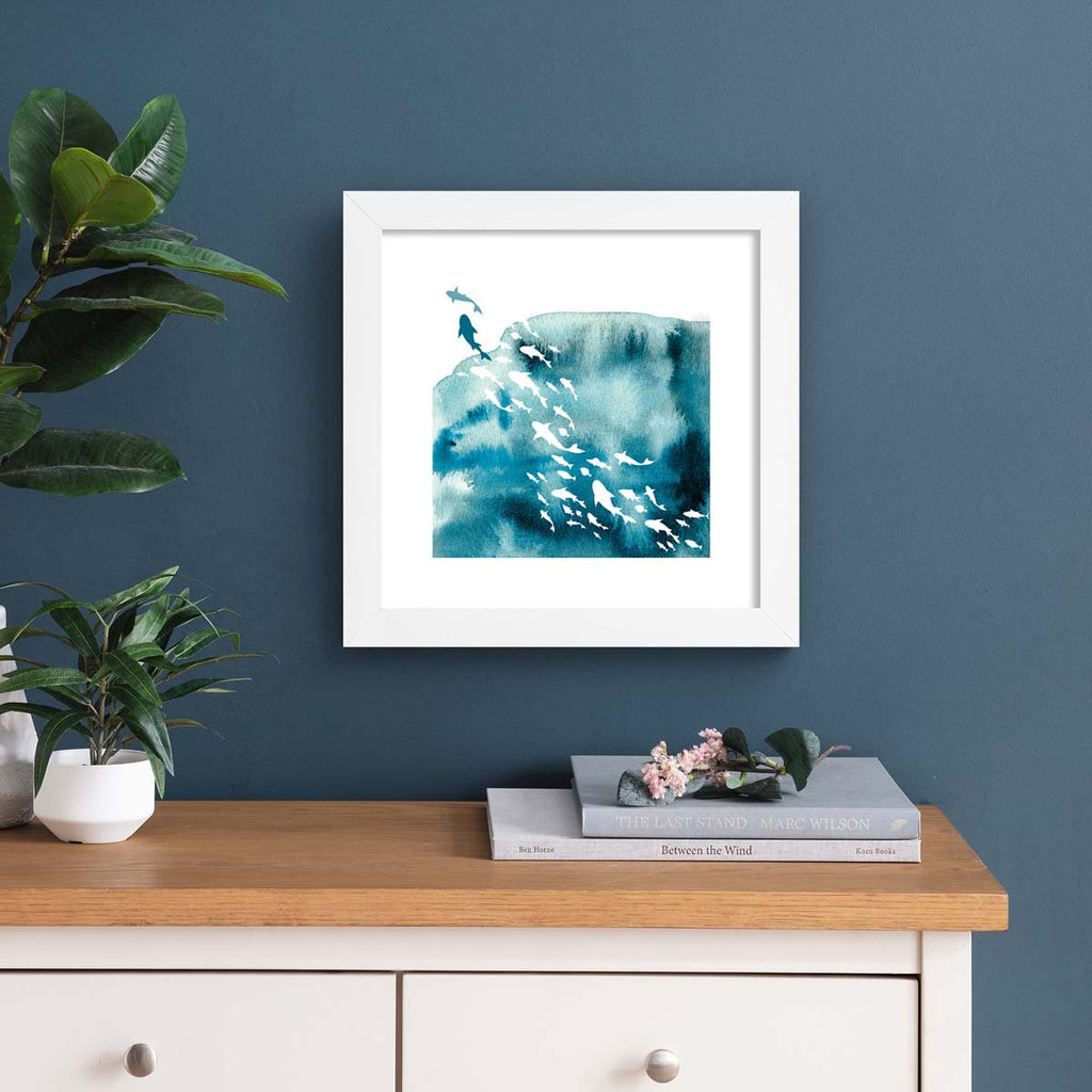 Vibrant watercolour art print featuring the silhouettes of fishes swimming in the ocean. Art print is hung up on a dark blue wall.